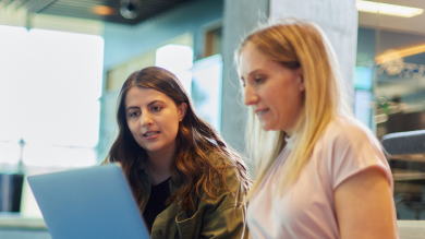 Two women sit together indoors, focused on a laptop screen in front of them, collaborating and discussing financial education in a modern office setting.