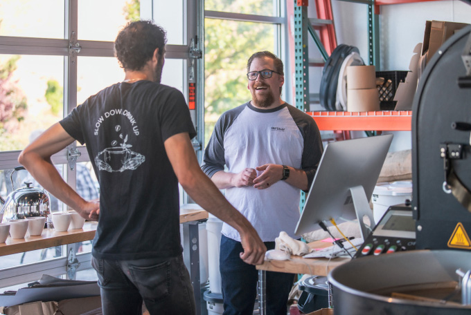 Two men are having a conversation in a modern workspace with shelves, a computer, and coffee cups; one man smiles at the camera while the other faces away, possibly discussing AI technology and public policy.