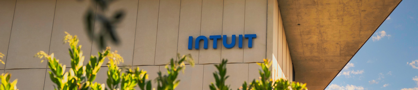 A building with the blue Intuit logo on its wall, symbolizing small business financial prosperity, is partially obscured by green foliage in the foreground, set against a blue sky with light clouds in the background.
