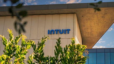 A modern building with the blue Intuit logo on its beige exterior wall, partially framed by green leaves and branches under a partly cloudy sky—a symbol of small business financial prosperity.