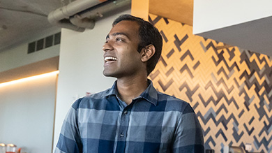 A man in a blue plaid shirt smiles and looks to the side in a modern room with patterned wall tiles, reflecting an atmosphere of Intuit accountability and transparency with its industrial-style ceiling.