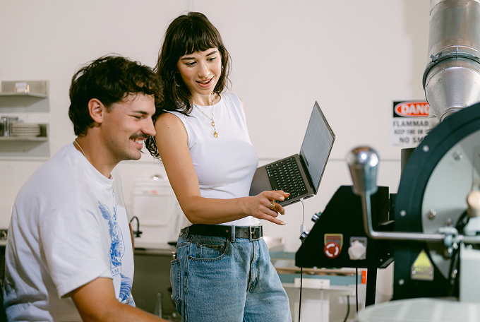 Two people in a lab setting, one sitting and smiling, the other standing with a laptop and pointing at equipment. A "Danger Flammable" sign is visible, highlighting the importance of small business financial tools for lab safety investments.