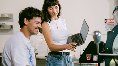 Two people in a lab setting interact; one sits smiling while the other stands holding a laptop, pointing at equipment. Safety signage and lab gear are visible in the background, highlighting teamwork much like small business financial tools foster collaboration.