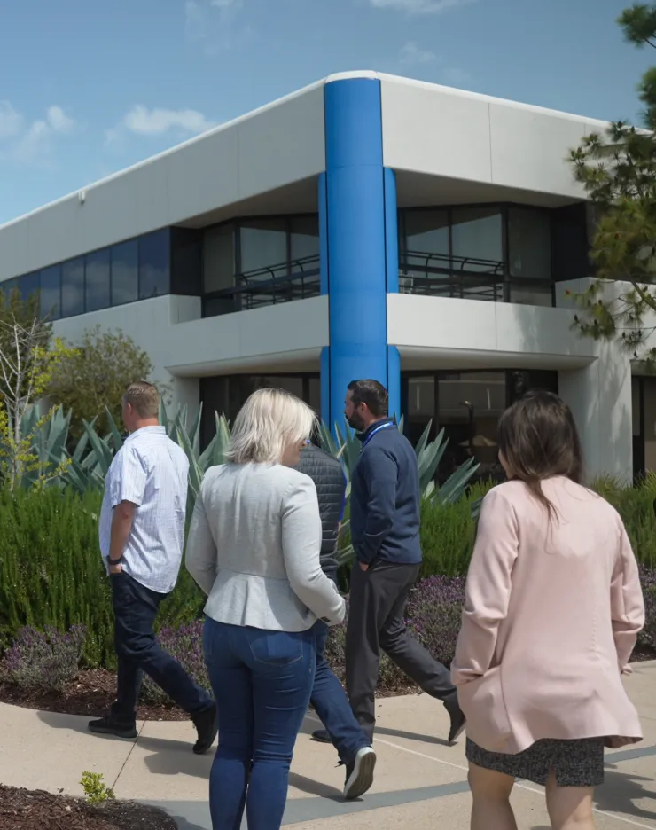 Five people walk together outside a modern office building with large windows and a blue column, discussing AI technology and public policy, surrounded by green landscaping and agave plants under a partly cloudy sky.