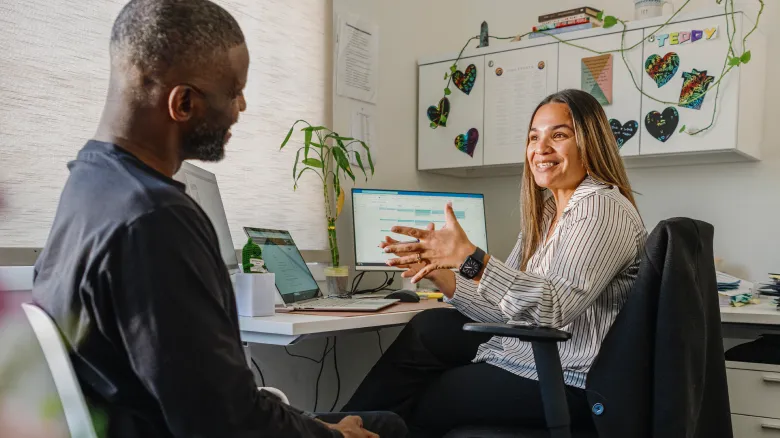 A woman sits at a desk, smiling and talking with a man seated across from her. In an office with plants, laptops, and colorful heart decorations, they discuss job creation through Intuit.