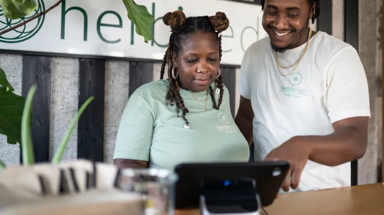 Two people wearing matching "herb n’ green" shirts stand at a counter, one exploring the best personal finance tool on a touchscreen device while the other smiles and points at the screen. A potted plant and a magazine sit in the foreground.