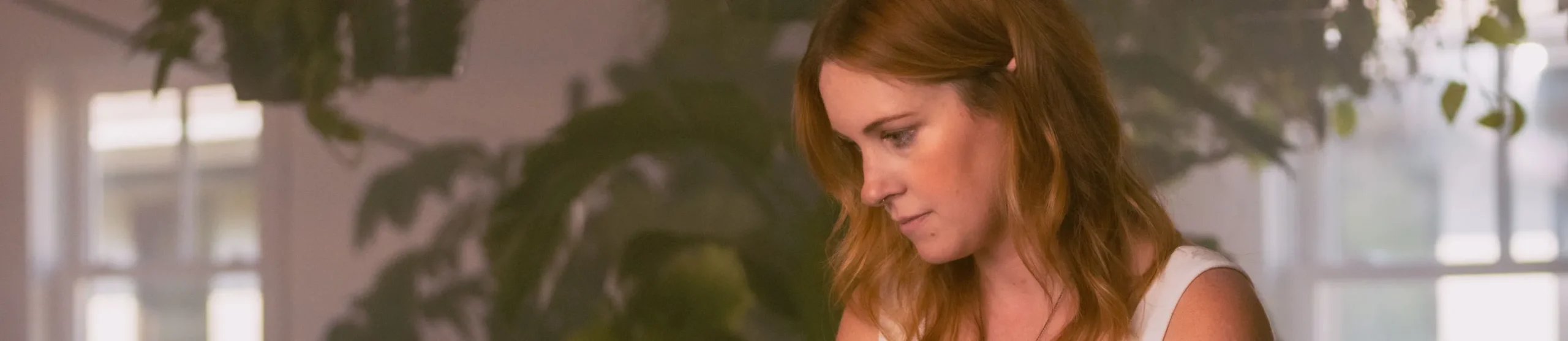 A woman with long red hair and a white top sits indoors, looking down and focused, surrounded by leafy plants and soft natural light—an atmosphere reflecting Intuit accountability and transparency.