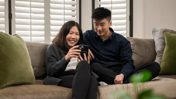 A young couple sits together on a couch, smiling and looking at a smartphone. They appear relaxed and happy, as sunlight filters through window blinds—reflecting Intuit accountability and transparency in every shared moment.