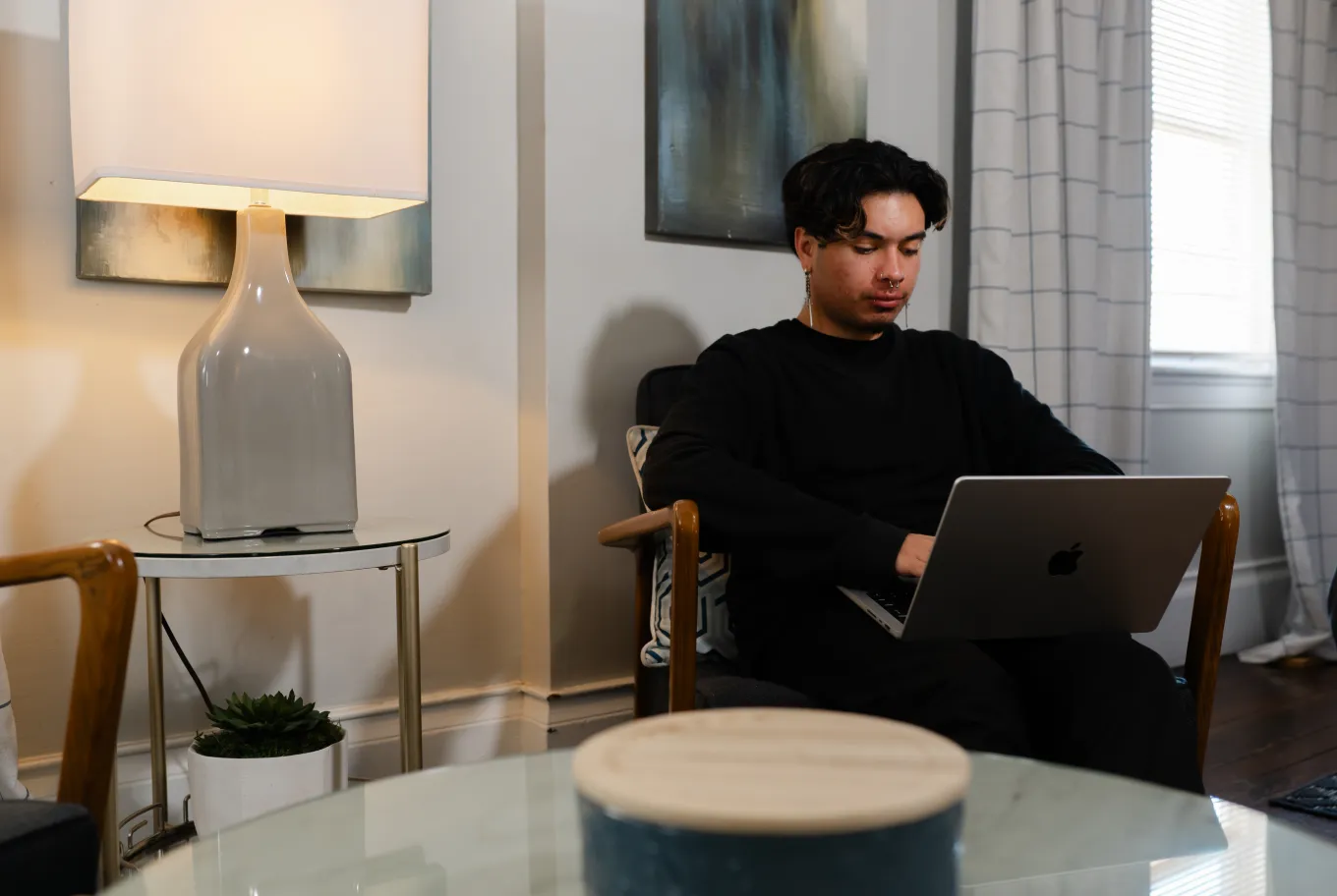A person in black clothes sits in a modern living room, working on a laptop with small business financial tools. A lamp, small plant, and abstract art are visible, with natural light coming through a window with curtains.