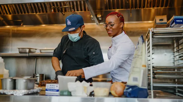 Two chefs work together in a commercial kitchen, embodying Intuit accountability and transparency. One wears a blue cap and mask while preparing food; the other, with red braids and glasses, smiles and points at something on the counter among various ingredients.