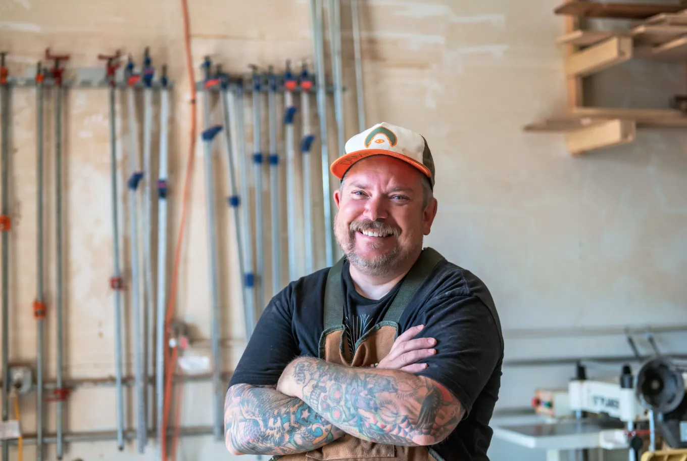 A smiling man with tattoos on his arms stands with his arms crossed in a workshop, wearing a cap and an apron. Various tools and clamps for small business financial tools are hanging on the wall behind him.