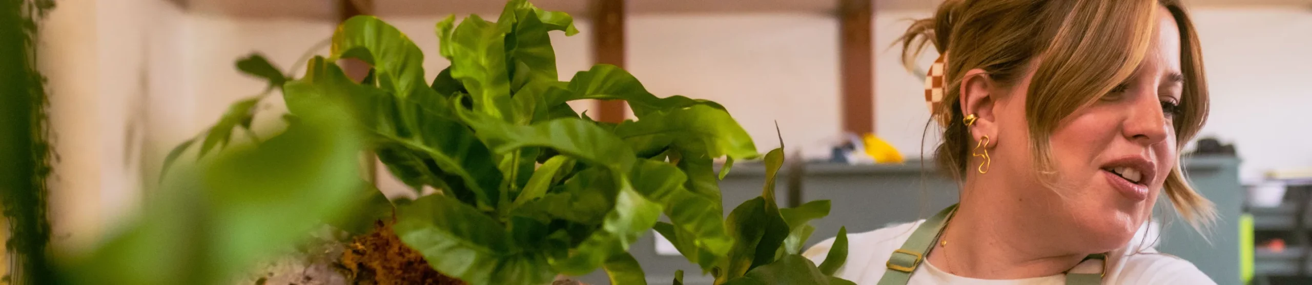 A woman with light brown hair and gold earrings smiles while holding a green potted plant indoors, embodying climate-positive technology solutions, with blurred shelves and wooden beams in the background.