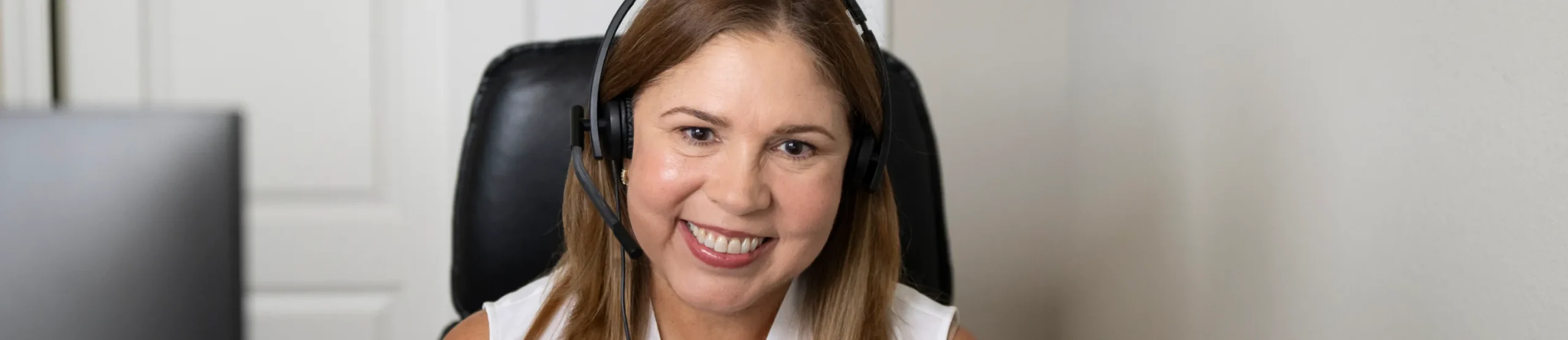 A woman with long brown hair, wearing a headset and white top, sits in a black chair and smiles at the camera in a well-lit indoor setting, ready to assist clients with personal finance and tax tools.