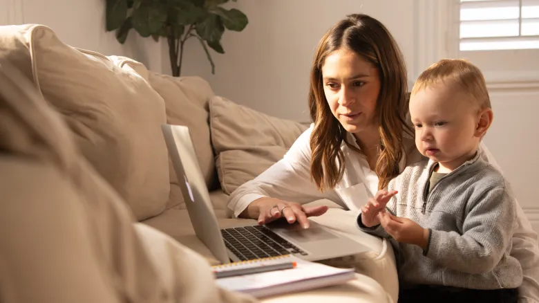 A woman sits on a couch using a laptop with a young child beside her. The child looks at the screen while the woman types, exploring AI technology. A notebook and pen are on the table in front of them. Natural light fills the room.