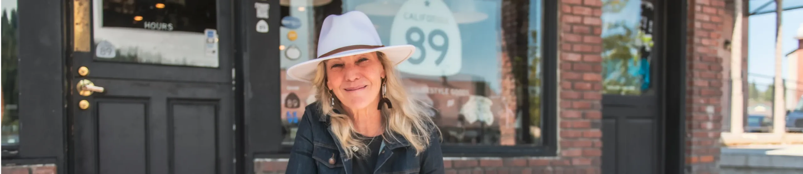 A smiling woman with long blonde hair wearing a white hat and dark jacket sits outside a brick building with large windows and a sign that reads "California 89," embodying the spirit of small business financial prosperity.
