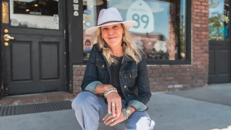 A woman with long blonde hair, wearing a white hat, denim jacket, and jeans, smiles while crouching on a sidewalk in front of a brick storefront—an inviting spot reflecting small business financial prosperity.