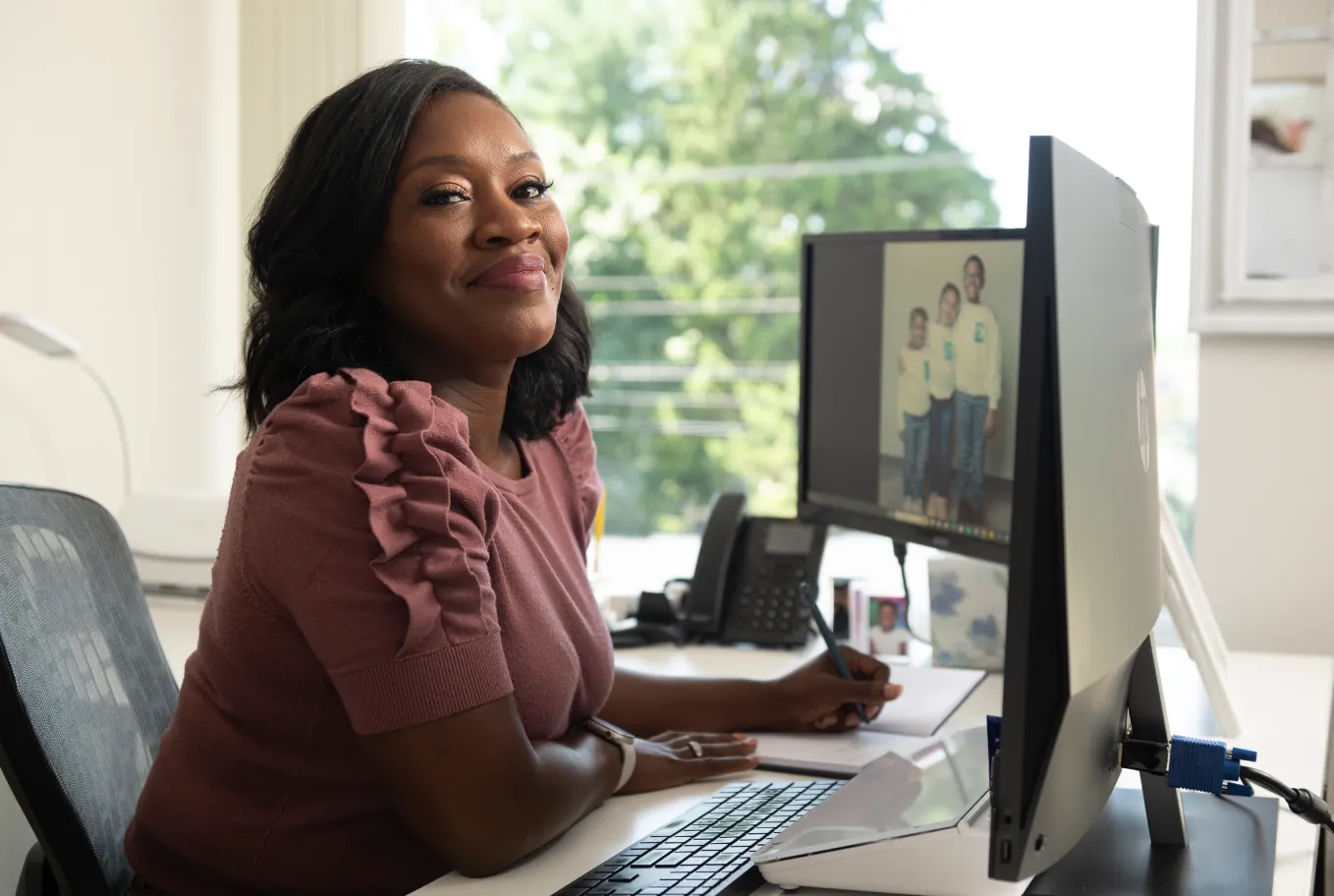 A woman sits at a desk, smiling at the camera, with one hand on a pen tablet and a family photo displayed on her computer monitor. The bright, modern office suggests she works in the field of AI technology and public policy.