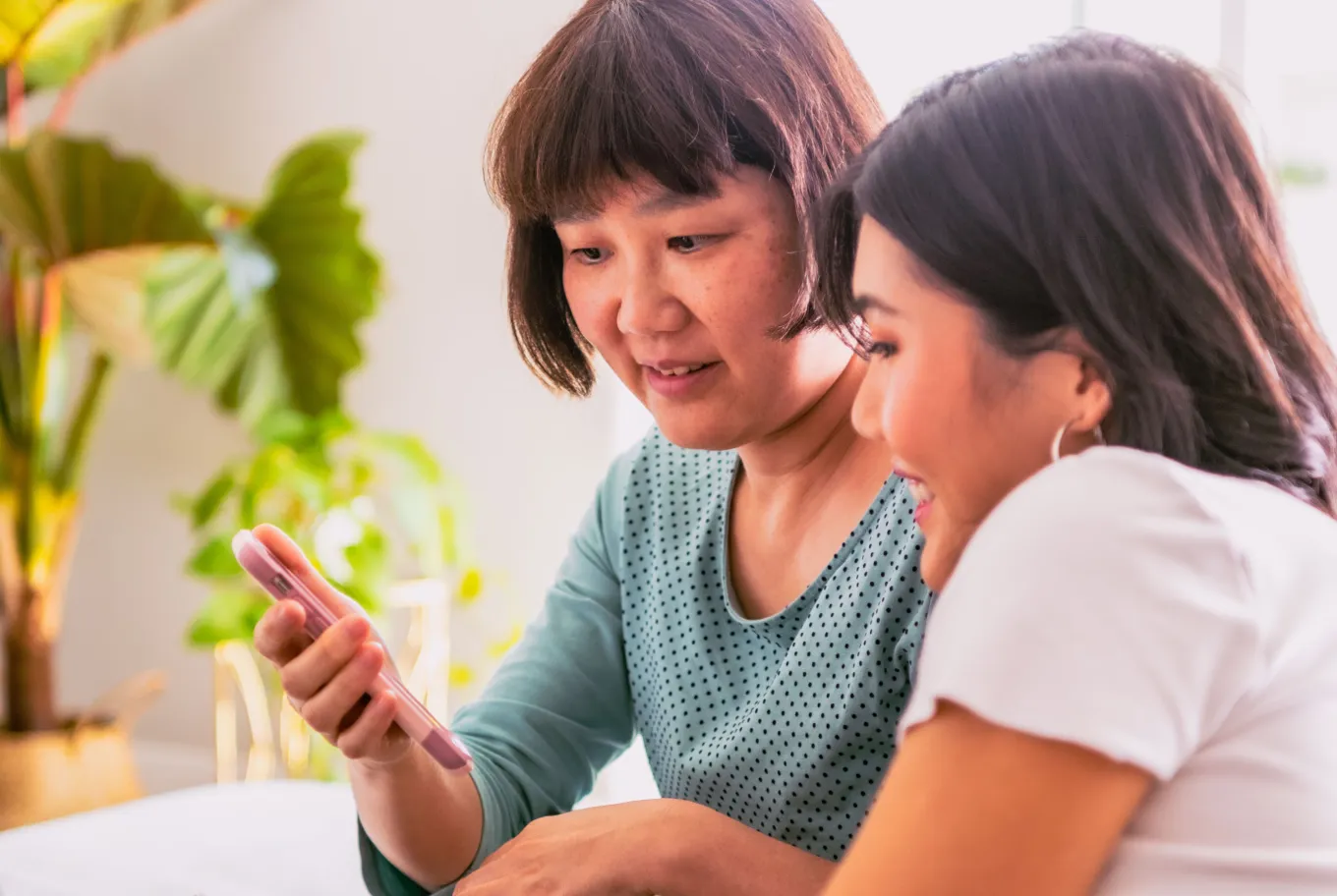 Two women sit closely together on a couch, smiling and looking at a smartphone showcasing climate-positive technology solutions. Green plants in the background and soft natural light fill the room.