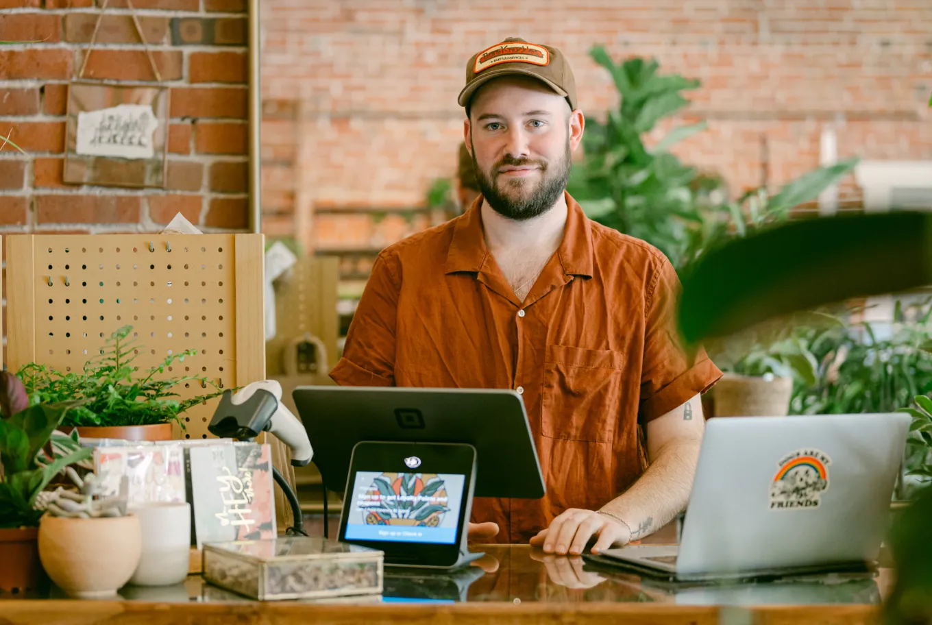 A man in a rust-colored shirt and hat stands behind a counter with a tablet, laptop, and plants, in a shop with brick walls and greenery, showcasing climate-positive technology solutions.