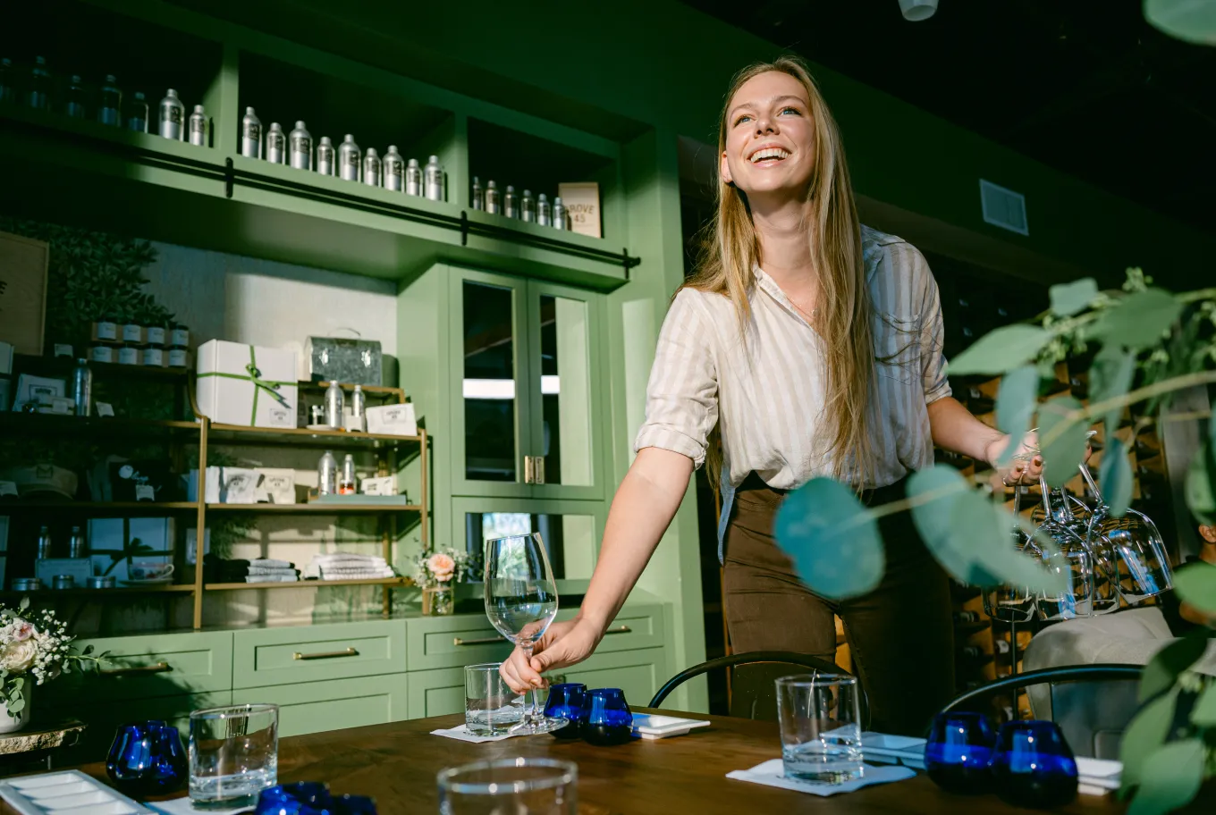 A smiling woman stands beside a dining table in a modern kitchen, holding a wine glass. The elegant setup reflects her small business financial prosperity, with blue candle holders, napkins, and sunlight streaming through the room.