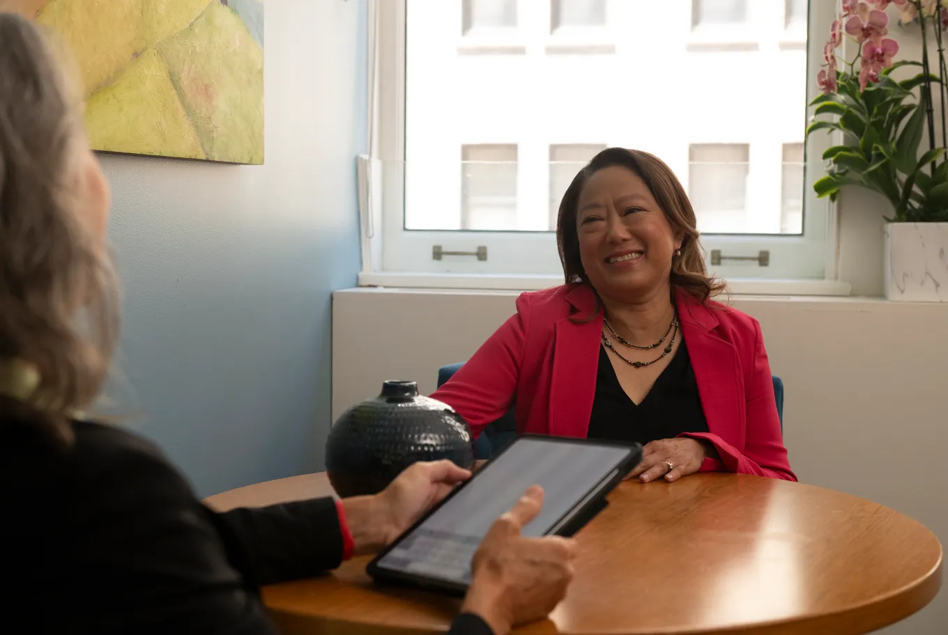 Two women sit at a round table in an office. One, smiling in a pink blazer, faces the camera while the other, with gray hair, holds a tablet displaying personal finance and tax tools on its screen.