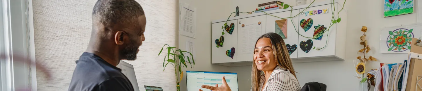 A woman sits at a desk, smiling and gesturing while talking with a man. Behind her are a computer monitor, plants, colorful artwork, and a whiteboard with notes about job creation through Intuit and other decorations.