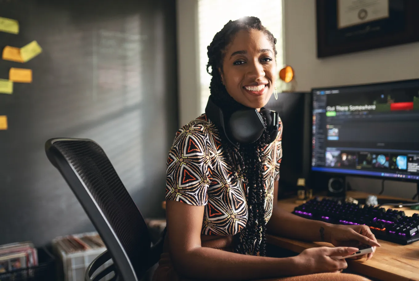 A young woman with braided hair and a patterned shirt sits at a computer desk, smiling, with headphones around her neck and the best personal finance tool open on her monitor. A keyboard and gaming controller are visible in the well-lit room.
