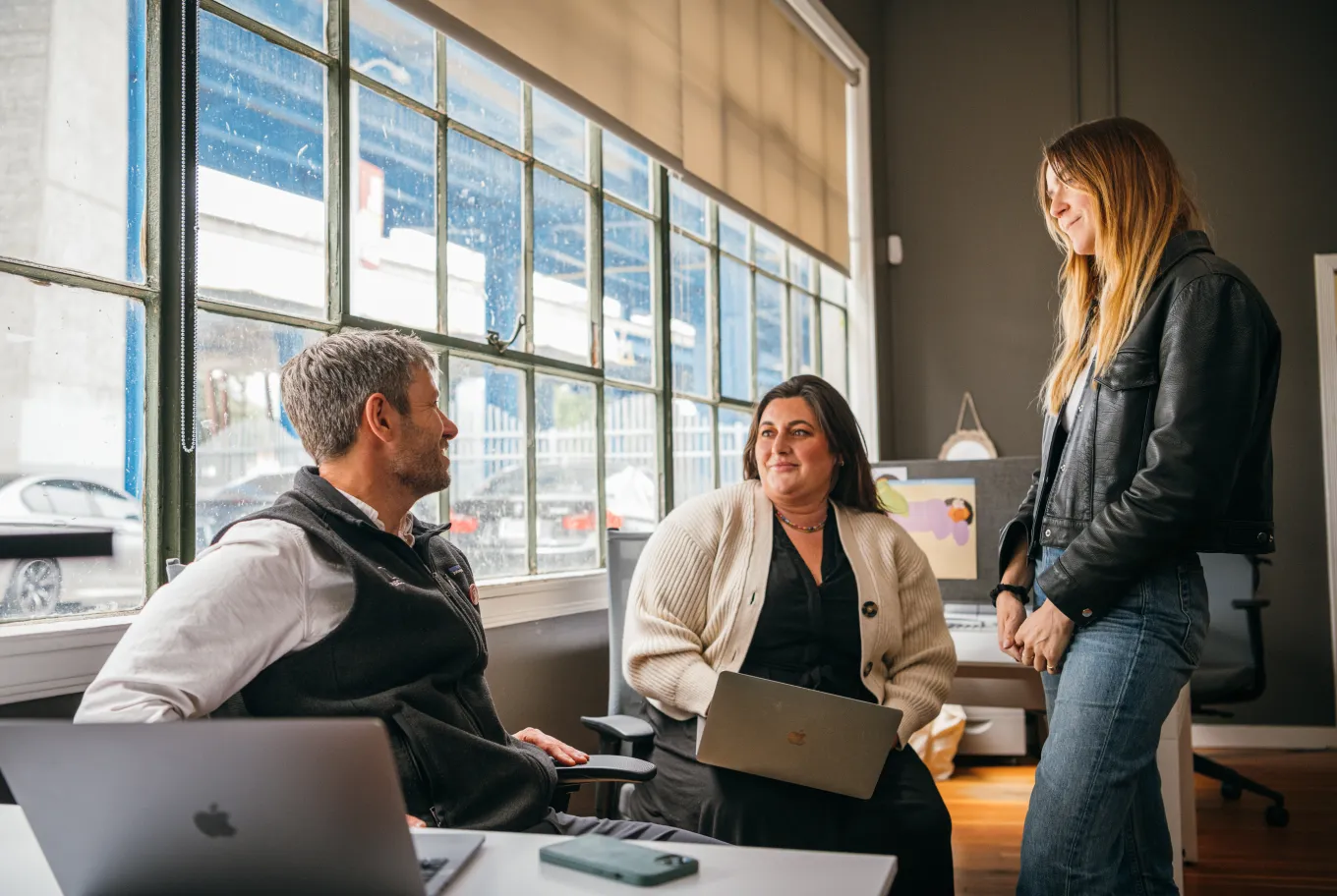 Three people are having a conversation in a modern office with large windows. Two are seated with laptops, while one stands and smiles, discussing job creation through Intuit. Natural light fills the room, and cars are visible outside.