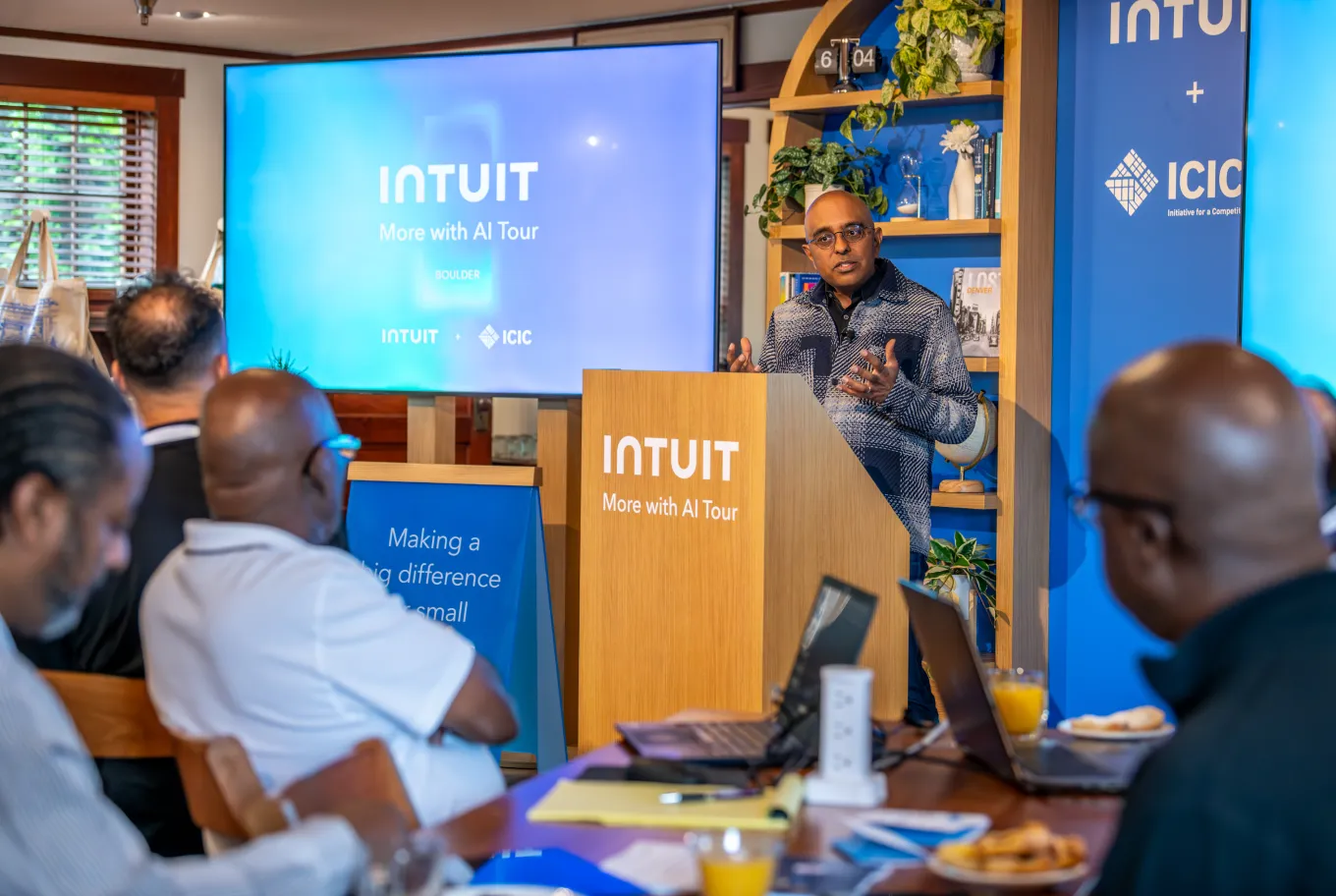 A man stands at a podium speaking to an audience during an Intuit AI Tour event, discussing AI technology and public policy. Large screens behind him display the Intuit logo as attendees with laptops and drinks listen attentively.