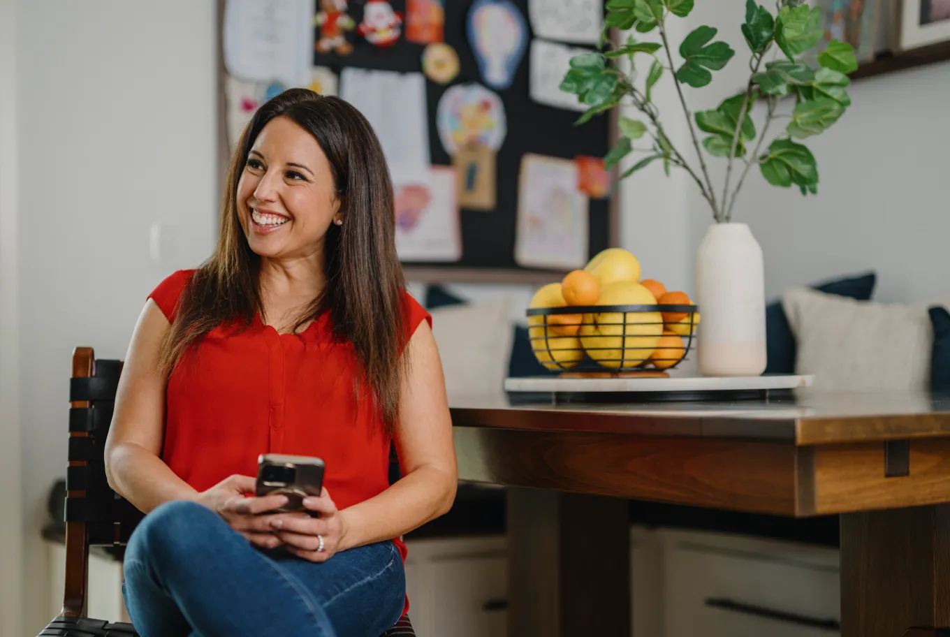 A woman in a red blouse sits on a chair, smiling and holding a smartphone. Behind her is a wooden table with a fruit bowl and vase, and a wall display with children’s artwork celebrating job creation through Intuit.