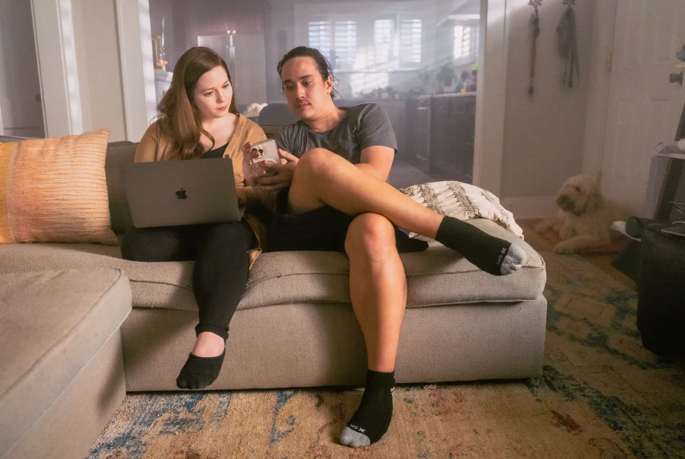 Two people sit on a couch; one explores AI technology on a laptop while the other shows something on a phone. A dog lies on the floor in the background, and natural light from a window softly illuminates the room.