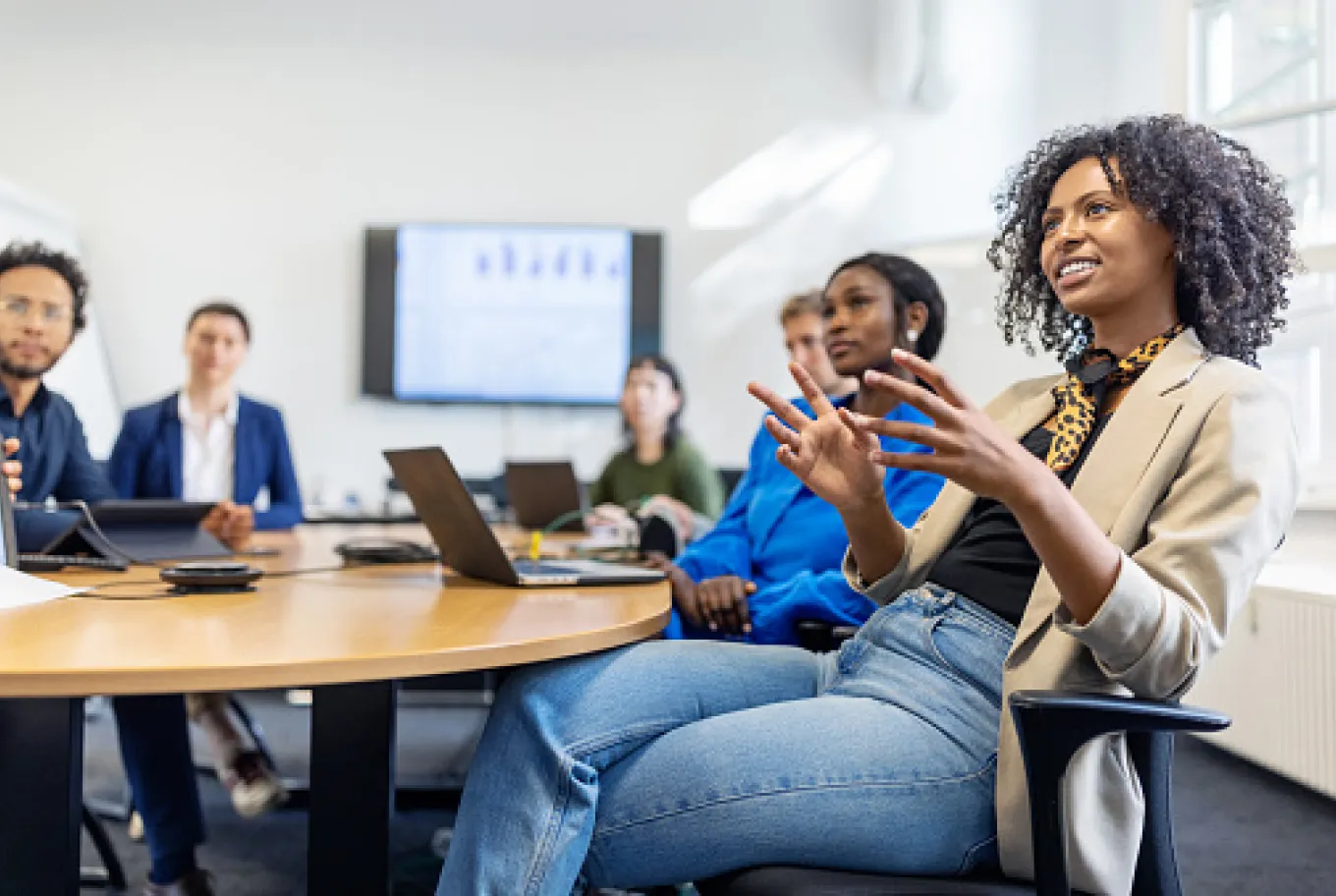 A group of people sit around a conference table in a meeting room, some with laptops. A woman in the foreground gestures as she speaks about small business financial prosperity, while others listen attentively. A large screen and window are visible behind them.
