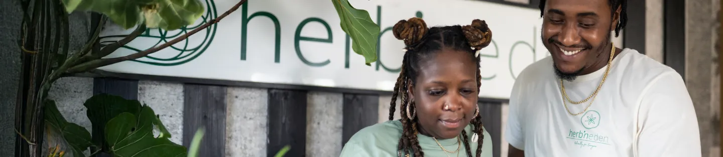 A smiling man and woman stand together indoors in front of a sign that reads “herb,” surrounded by green plants. Both are wearing light-colored shirts with matching logos, showcasing their pride in creating the best personal finance tool.