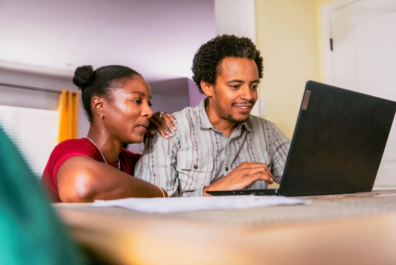 A couple sits together at a table, smiling and looking at a laptop screen as they explore small business financial tools, working side by side in a brightly lit room with one person’s arm around the other.
