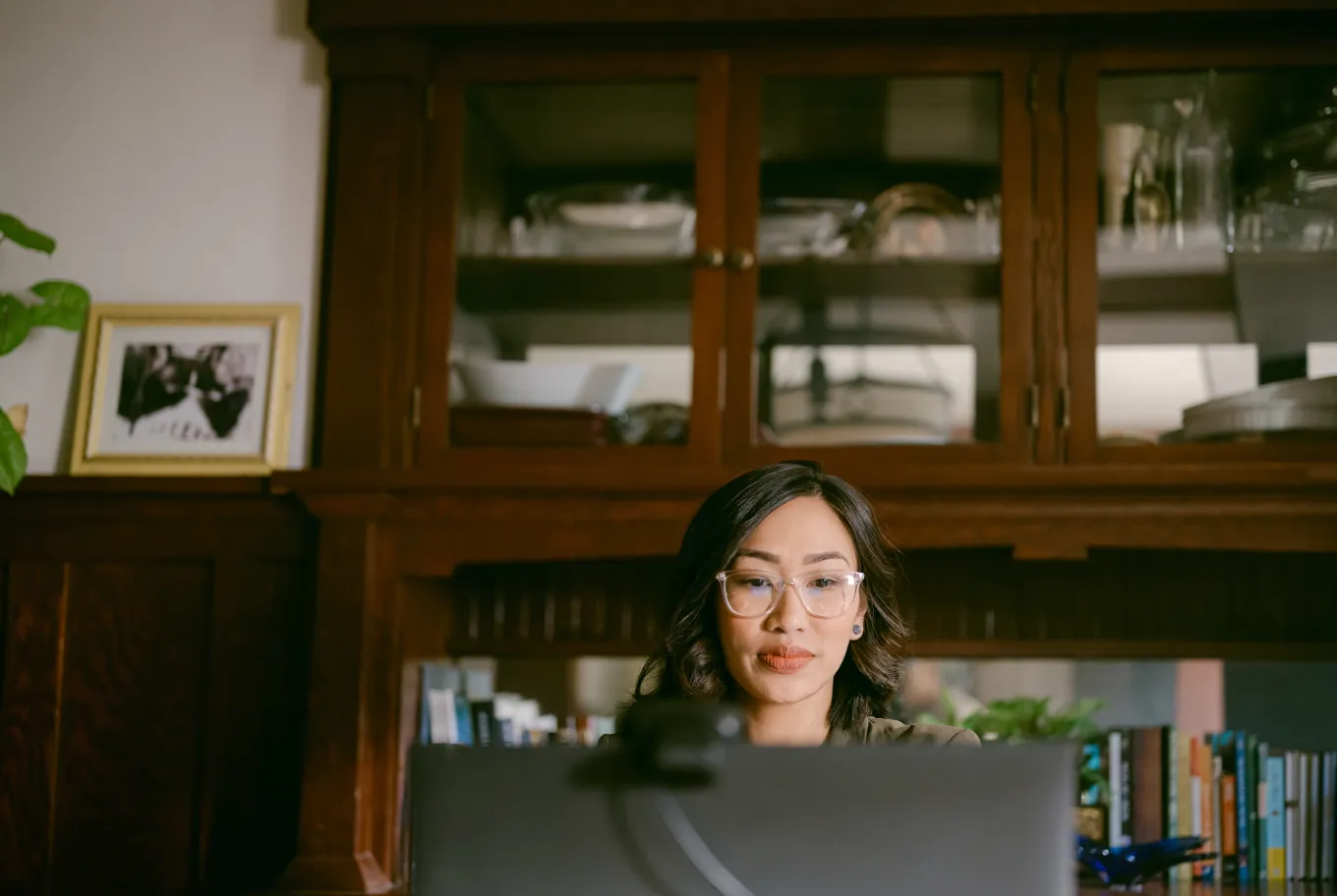 A woman wearing glasses sits at a desk, using personal finance and tax tools on her computer. Behind her is a wooden cabinet with glass doors, shelves lined with books, and a framed photograph.