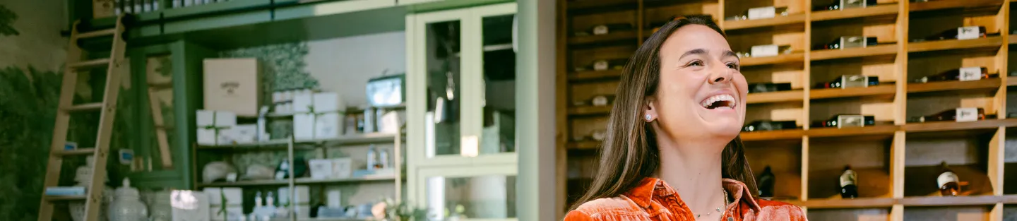 A smiling woman with long dark hair, wearing an orange shirt, stands indoors next to wooden shelves filled with bottles. The background shows a green cabinet and shop items, highlighting the charm of small business financial tools in a cozy setting.