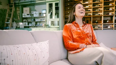 A woman in an orange blouse sits on a light-colored couch, smiling and laughing. Behind her are shelves with bottles and jars, reflecting the inviting charm of a small business that may benefit from financial tools in this cozy, well-lit space.