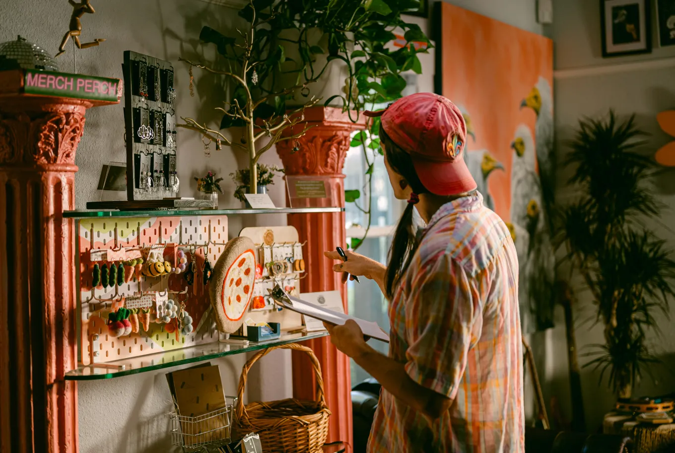 A person in a red cap and plaid shirt browses colorful keychains and small items on glass shelves in a cozy, plant-filled shop that highlights climate-positive technology solutions.