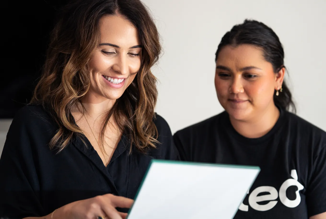 Two women smiling and looking at a document or screen together, discussing personal finance and tax tools. One woman points at the document, and both appear engaged and happy. The background is plain and uncluttered.