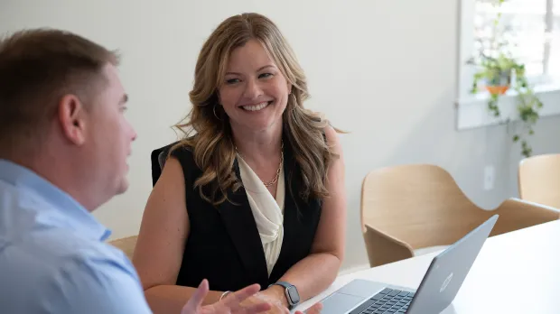 A woman smiles while talking with a man at a table in a bright office, reflecting Intuit accountability and transparency. Both are seated near a laptop, and the room features light-colored walls and modern chairs.