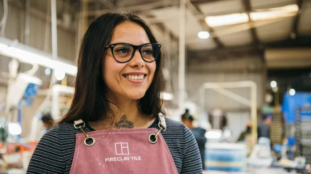 A woman with long dark hair and glasses smiles in a pink “Fireclay Tile” apron, embodying Intuit accountability and transparency inside a well-lit workshop where equipment and people work in the blurred background.