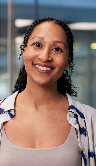 A woman with curly dark hair smiles at the camera. She is wearing a white and blue patterned shirt over a beige top, and stands in front of a blurred office background.