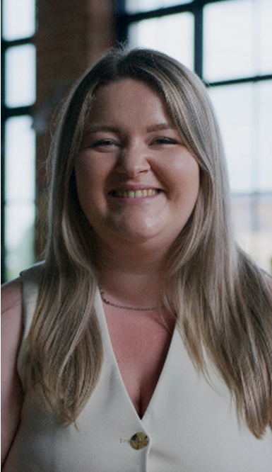 A woman with long blonde hair, wearing a light-colored sleeveless top, smiles while standing indoors in front of large windows with a blurred background.