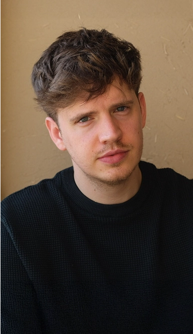 A young man with short, wavy brown hair and light facial hair wears a black sweater and looks directly at the camera against a beige background.