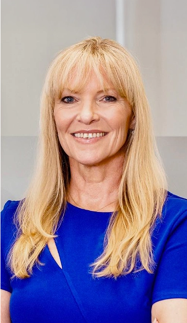 A woman with long blonde hair and bangs is smiling and wearing a bright blue short-sleeved dress, standing against a neutral indoor background.