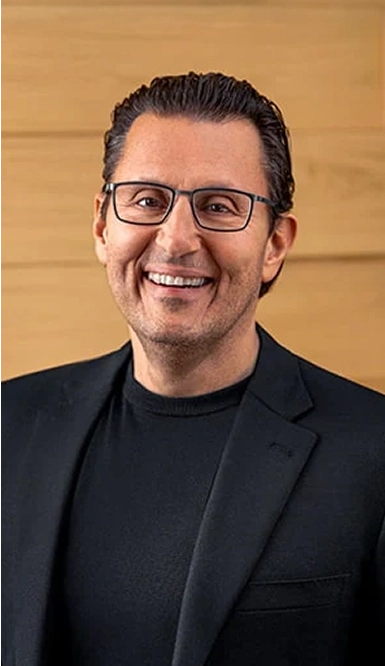 A man with short dark hair, wearing glasses, a black blazer, and a black shirt smiles while standing in front of a light wooden wall.