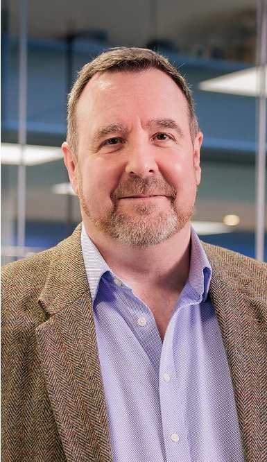 A middle-aged man with short brown hair and a trimmed beard, wearing a light blue button-up shirt and a brown tweed jacket, smiles slightly while standing indoors in a modern office setting.