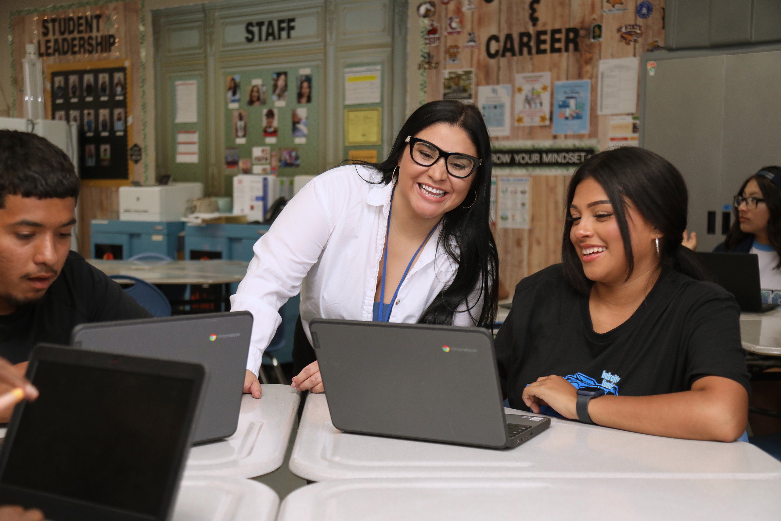 A teacher smiles while assisting a student working on financial education tasks on a laptop in a classroom. Another student sits nearby, also on a laptop. Classroom walls display posters about leadership, staff, and careers.