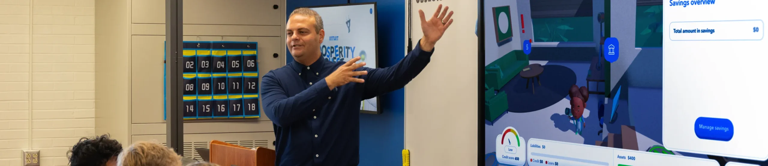 A man gestures while speaking in front of a classroom about financial education, with students seated and a large digital screen showing an animated game interface behind him.